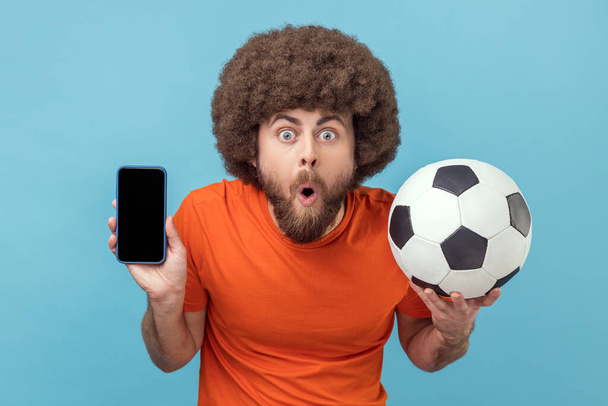 Portrait of shocked man with Afro hairstyle wearing T-shirt standing with soccer ball and showing cell phone with empty display for advertisement. Indoor studio shot isolated on blue background. - Foto, Bild