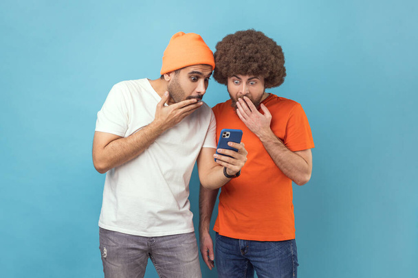 Portrait of two attractive shocked young adult hipster men standing, using smartphone, reading breaking news in social networks, covering open mouth. Indoor studio shot isolated on blue background. - Foto, imagen