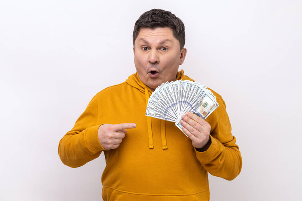 Shocked man pointing at big fan of dollars banknotes with finger, being astonished by winning lottery, looking at camera, wearing urban style hoodie. Indoor studio shot isolated on white background. - Photo, image