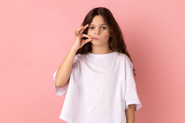 Portrait of charming little girl wearing white T-shirt with mystery look making zip gesture to close mouth, keeping secret, zipping lips. Indoor studio shot isolated on pink background. - Фото, изображение