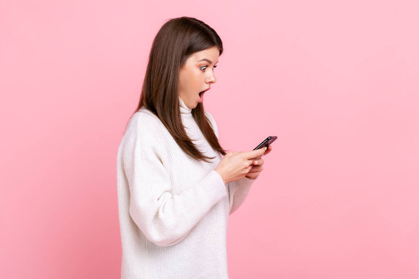 Side view portrait of shocked blogger woman standing, use smartphone with amazed face, opened mouth, wearing white casual style sweater. Indoor studio shot isolated on pink background. - Фото, изображение