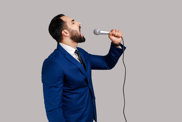 Side view of bearded man singing songs, holding microphone, singer making performance, keeps eyes closed, wearing official style suit. Indoor studio shot isolated on gray background. - Photo, Image