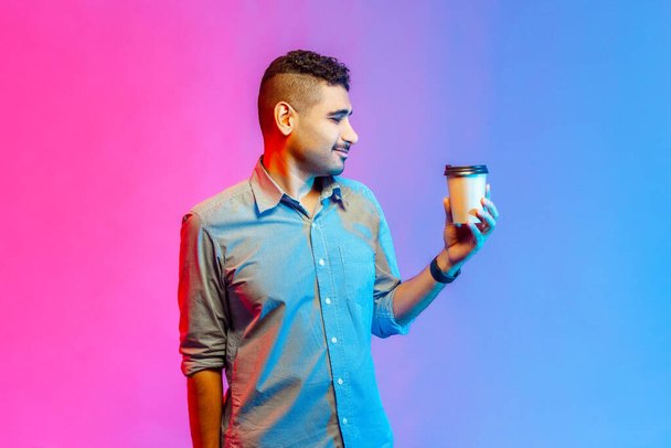 Portrait of handsome young adult man in shirt holding paper cup with morning coffee, looking at hot beverage, enjoying. Indoor studio shot isolated on colorful neon light background. - Fotografie, Obrázek