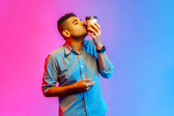 Portrait of handsome young adult man in shirt kissing disposable cup of take away coffee, enjoying beverage, needs energy. Indoor studio shot isolated on colorful neon light background. - Fotografie, Obrázek
