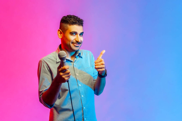 Portrait of attractive man in shirt posing with microphone in hands, offers mic, journalist asking questions, showing thumb up. Indoor studio shot isolated on colorful neon light background. - Foto, Imagen