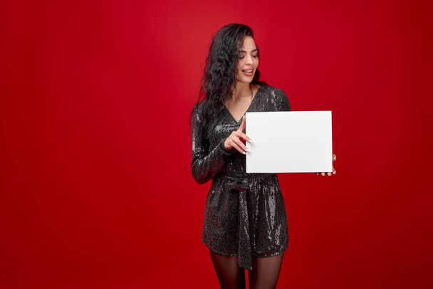 A young girl in a black short dress on a red background holds an empty white box. A beautiful brunette in a black bodysuit holds a white box with space for text or logo. Discount advertising. - Fotó, kép