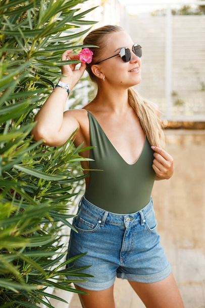 Portrait of a pretty blonde woman in a top and denim shorts, summer vacation, a girl with a pink flower near her face - Photo, image