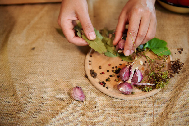 Still life. Housewifes hands, holding fragrant culinary herbs above a chopping board with laid out garlic cloves, fresh dill leaves, dry bay leaves and scattered peppercorns on a linen tablecloth - Foto, afbeelding