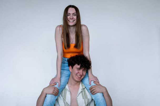 Cheerful young couple in bright summer clothes, gray studio background, copy space. Smiling girl sitting on shoulders of happy guy in unbuttoned shirt. Casual women and men wear for vacation. - Photo, image