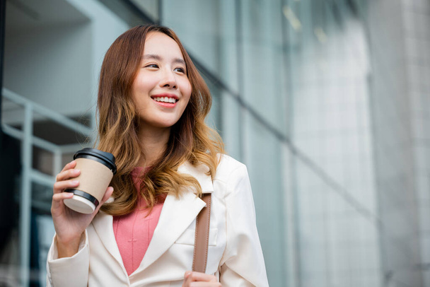 Happy business woman smiling hold paper cup of hot drink outdoor walking on street, Asian businesswoman holding coffee cup takeaway going to work she walking near her office building outside - Foto, afbeelding
