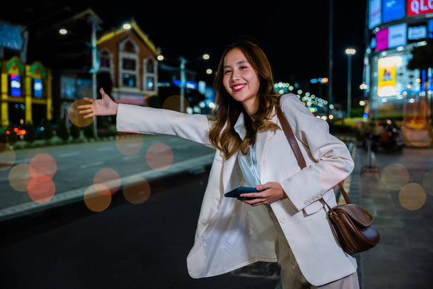 Asian businesswoman standing hail waving hand taxi on road in busy city street at night, beautiful woman smiling using mobile phone application hailing with hand up calling cab after late work - Zdjęcie, obraz