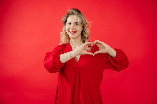 Portrait of happy attractive young woman in beautiful red look with perfect bright makeup isolated on red background. Love expression, making heart shape by hands. Fashion and beauty industry. - Foto, imagen