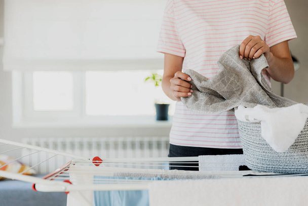Γυναίκα κρέμονται καθαρά βρεγμένα ρούχα πλυντήριο σε σχάρα στεγνώματος στο σπίτι στο δωμάτιο. Γυναίκα νοικοκυρά χέρια closeup εκμετάλλευση, διάδοση πλυντήριο από το καλάθι. Δουλειές. Κορίτσι κρατά διπλωμένες πετσέτες μπάνιου στο χέρι - Φωτογραφία, εικόνα