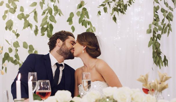 When they kiss, they do it with passion. an affectionate young newlywed couple kissing while sitting at their wedding reception - Φωτογραφία, εικόνα
