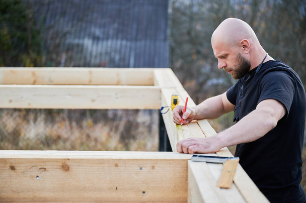 Man worker building wooden frame house on pile foundation. Bald carpenter using tape measure for measuring wooden planks and making marks with pencil. Carpentry concept. - Fotoğraf, Görsel