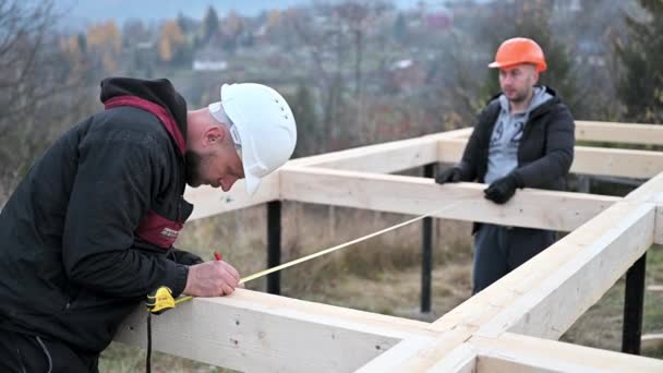 Men workers building wooden frame house on pile foundation. Carpenters using tape measure for measuring wooden planks and making marks with pencil. Carpentry concept. - Video