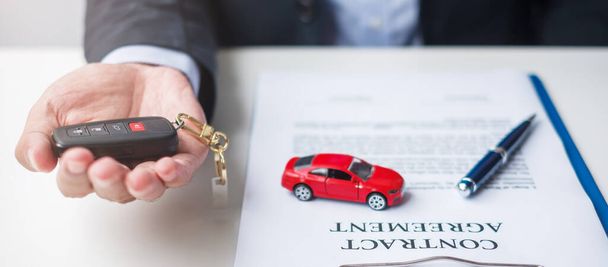 Επιχειρηματίας χέρι κρατώντας κόκκινο αυτοκίνητο παιχνίδι με το όχημα keyless, στυλό και το έγγραφο της σύμβασης. έννοιες συμβάσεων αγοράς και πώλησης, ασφάλισης, μίσθωσης και σύμβασης - Φωτογραφία, εικόνα