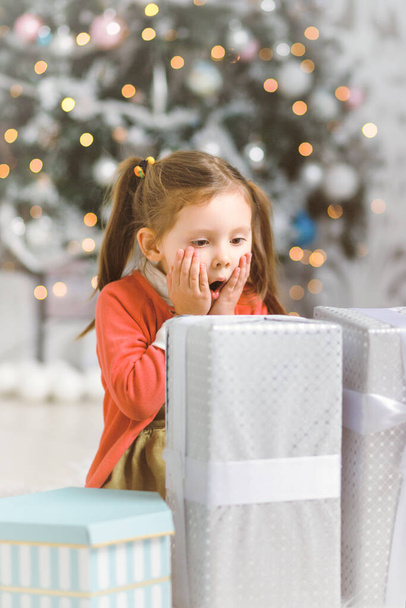Emotional little girl with New Year gifts in white interior with christmas tree. - Foto, Imagen