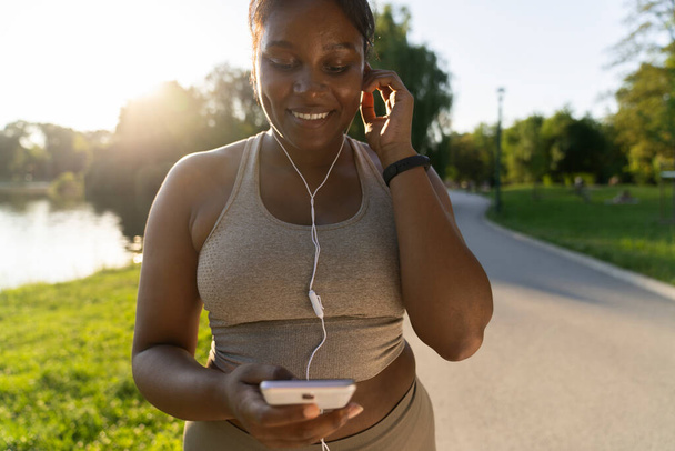 African American woman using mobile phone and earphones before exercising at the park in a  summer day  - 写真・画像