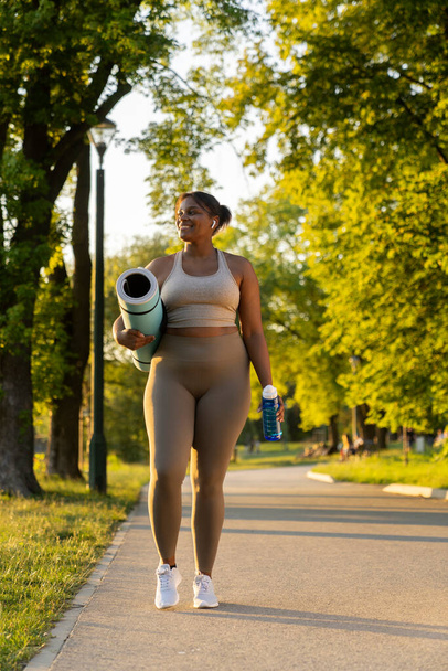 Plus sized African American woman walking with exercise mat and water through the park in a summer day - Fotografie, Obrázek