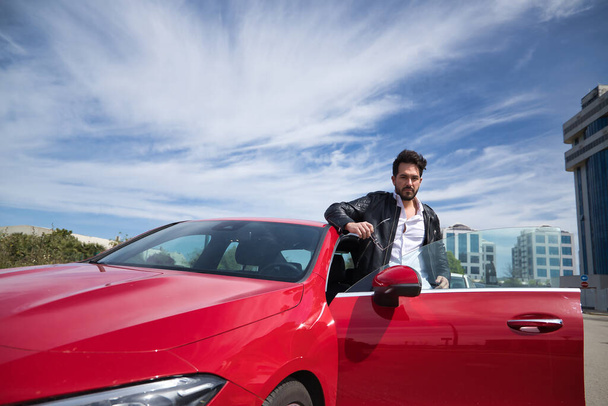 Handsome young man with beard, sunglasses, leather jacket and white shirt, leaning on the roof of his red sports car, looking at the camera seriously. Concept beauty, fashion, luxury, motor, sports. - Φωτογραφία, εικόνα