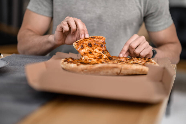 Eating pizza. Hands of man sitting at table at home taking slice of delicious pizza without face - Foto, Imagem