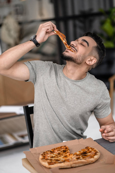 Pleasure. Young dark haired man with closed eyes biting pizza holding piece above face sitting at table in apartment - Foto, Imagem