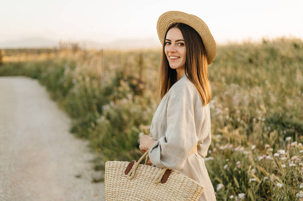 Portrait of a happy woman walking with a straw bag through the field at sunset, enjoying nature in summer. Concept of wellness and carefree lifestyle in nature - Zdjęcie, obraz