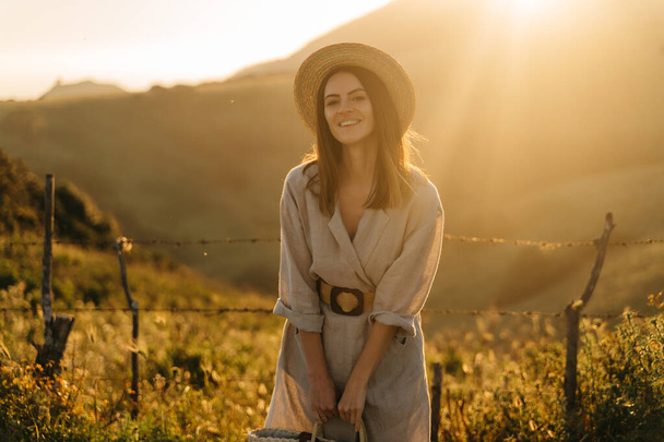 Portrait of a happy woman walking with a straw bag through the field at sunset, enjoying nature in summer. Concept of wellness and carefree lifestyle in nature - Photo, Image