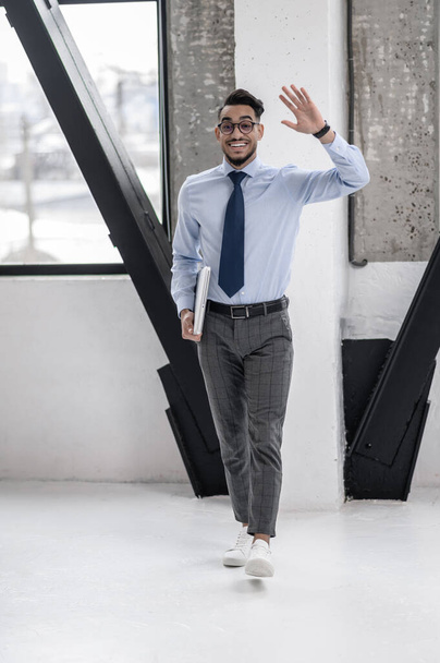 All excellent. Happy business young man in tie and glasses holding laptop in greeting gesturing energetically walking towards camera indoors - Foto, Imagen