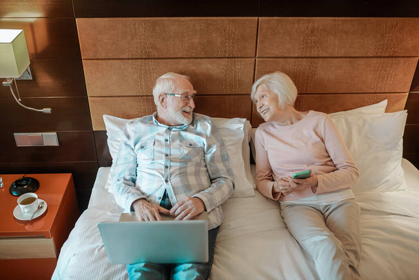 Resting in a hotel. Senior couple lying on bed in a hotel room and watching something online - Fotografie, Obrázek