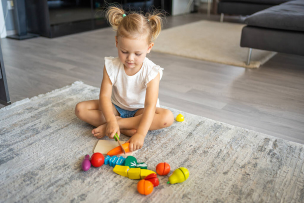 Cute little toddler girl playing at home with eco wooden toys. Happy healthy excited child cutting vegetables and fruits with toy knife. The child playing educational games. High quality photo - Фото, изображение