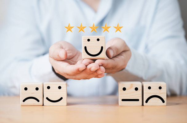 Businessman holding smile face on wooden cube block with other face emotion for excellent evaluation and customer satisfaction concept. - 写真・画像