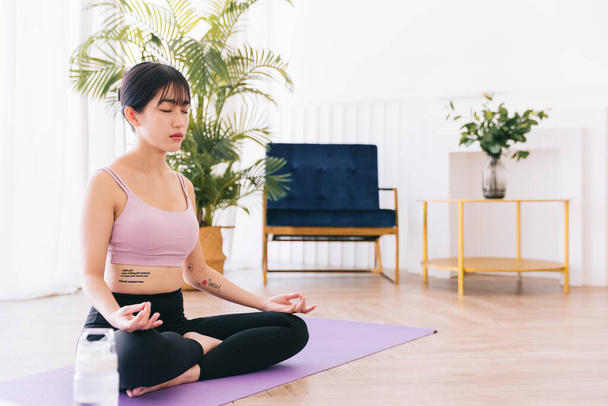 Giovane attraente donna asiatica sportiva che pratica yoga su un tappetino yoga, facendo esercizio Ardha Padmasana, meditando in posa Half Lotus, indoor lavorando a casa, indossando abbigliamento sportivo. - Foto, immagini