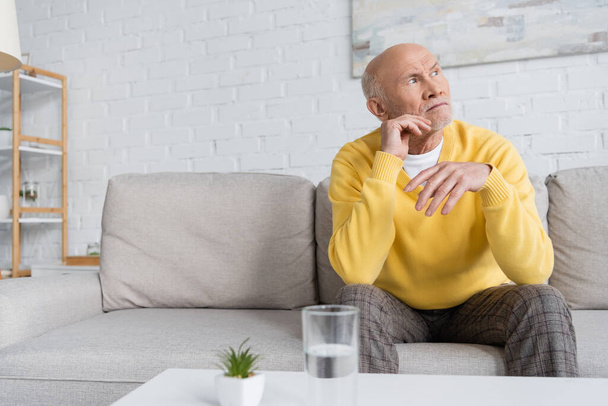 Elderly man sitting near blurred glass of water in living room at home  - Fotoğraf, Görsel