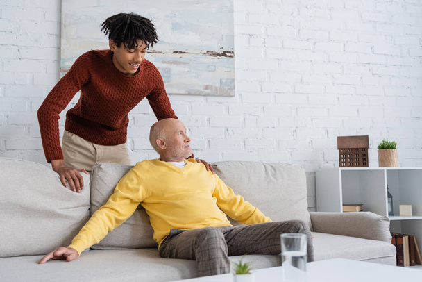 Smiling african american grandson standing near senior granddad on couch in living room  - Photo, Image
