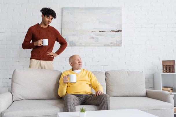 African american grandson holding cup near grandpa sitting on couch at home  - Valokuva, kuva