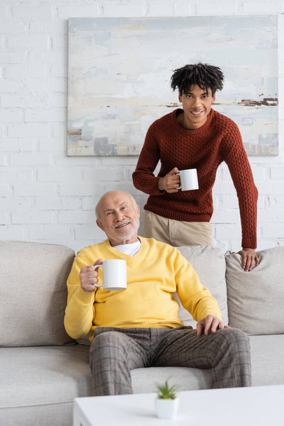 Smiling multiethnic grandfather and grandson holding cups and looking at camera at home  - Фото, зображення
