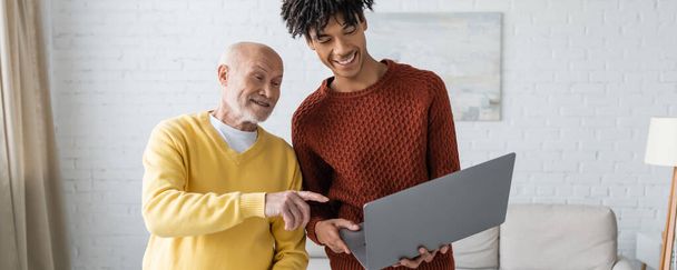 Positive pensioner pointing at laptop near african american grandson at home, banner  - Foto, immagini