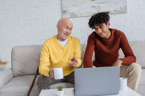 Cheerful pensioner pointing at laptop and holding cup near african american grandson at home  - Foto, Imagem