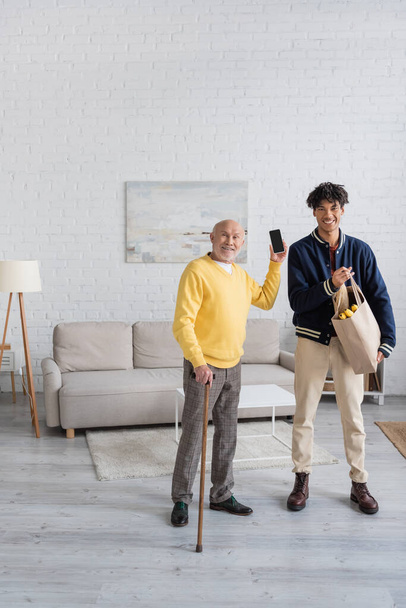 Cheerful pensioner holding smartphone near african american grandson with food at home  - Foto, Bild