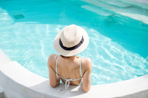 Ευτυχισμένη γυναίκα σε λευκό μαγιό που κολυμπά σε πολυτελές ξενοδοχείο πισίνας, νεαρή γυναίκα με καπέλο απολαμβάνουν σε τροπικό θέρετρο. Χαλαρωτικό, καλοκαιρινά ταξίδια, διακοπές, διακοπές και Σαββατοκύριακο έννοια - Φωτογραφία, εικόνα