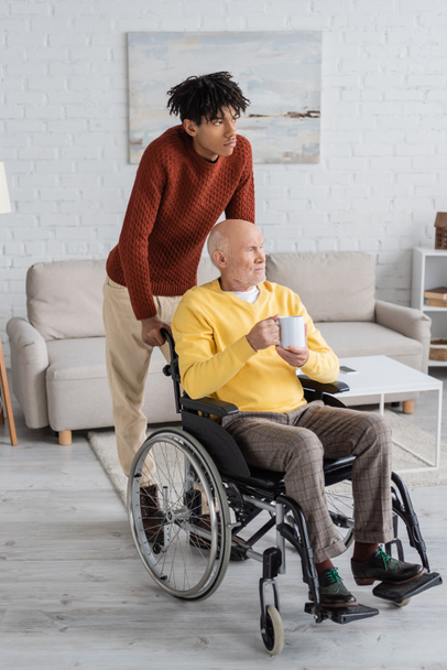 African american man standing near granddad holding cup in wheelchair at home  - Foto, Bild