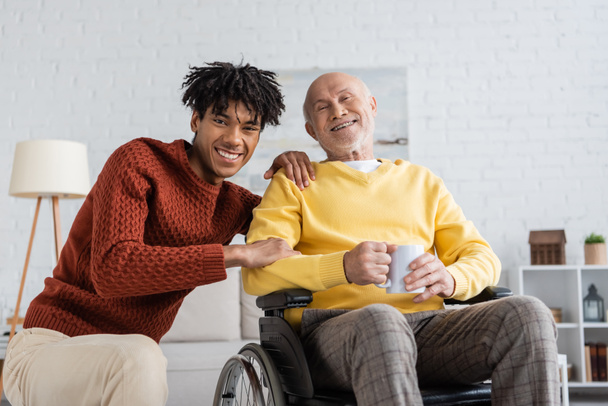 Smiling african american grandson hugging granddad with cup in wheelchair at home  - Fotó, kép