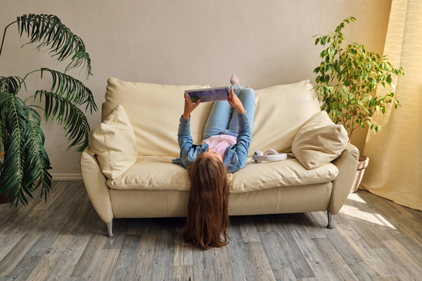 маленькая девочка лежит вверх ногами на диване и играет с планшетом - Фото, изображение