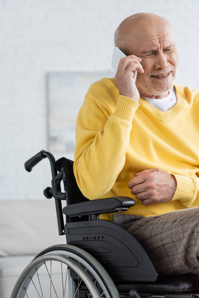 Disappointed elderly man talking on smartphone while sitting in wheelchair at home  - Foto, Imagem