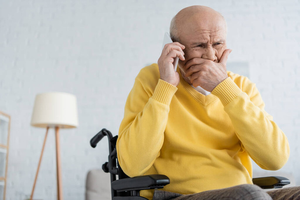 Upset senior man talking on smartphone while sitting in wheelchair at home  - Photo, Image