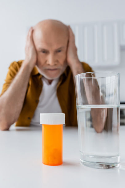 Glass of water and pills near blurred elderly man at home  - Фото, зображення