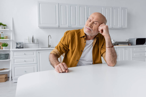 Sad senior man sitting at white table in kitchen  - Fotoğraf, Görsel
