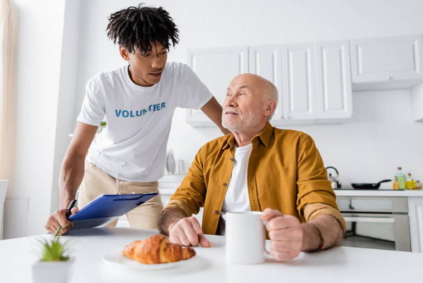 African american volunteer holding clipboard near elderly man with cup and croissant at home  - Photo, Image